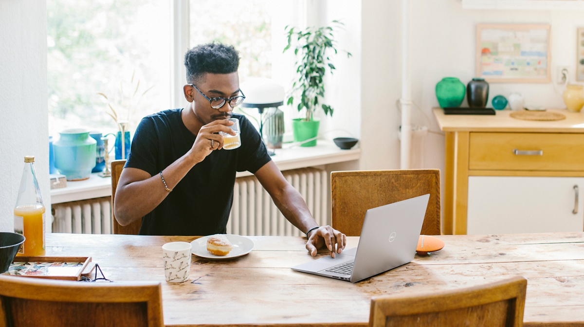 Photo d’un homme tenant un verre de jus dans sa main droite et utilisant un ordinateur portable Dell Inspiron 16 5620 placé sur une table devant lui.