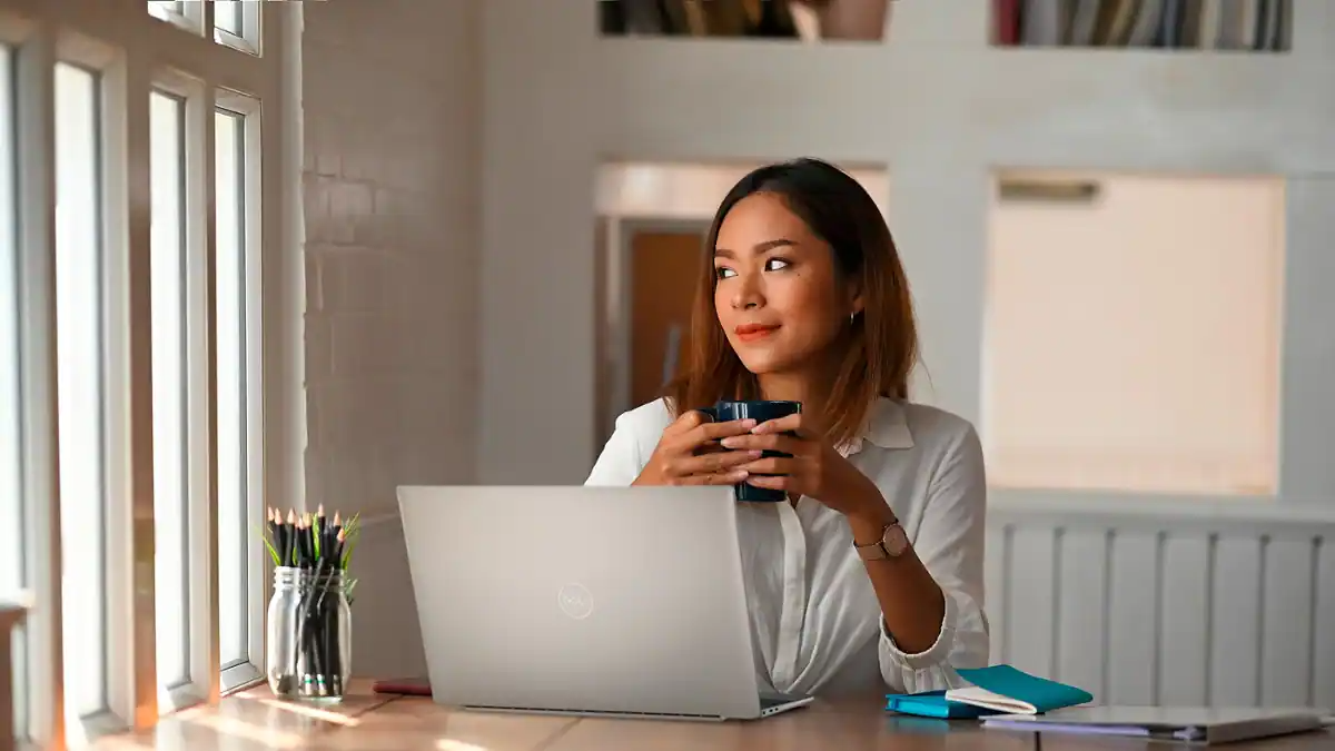 Photo d’une femme ayant une tasse dans les mains et utilisant un ordinateur portable Dell XPS 13 9320 posé sur une table devant elle.